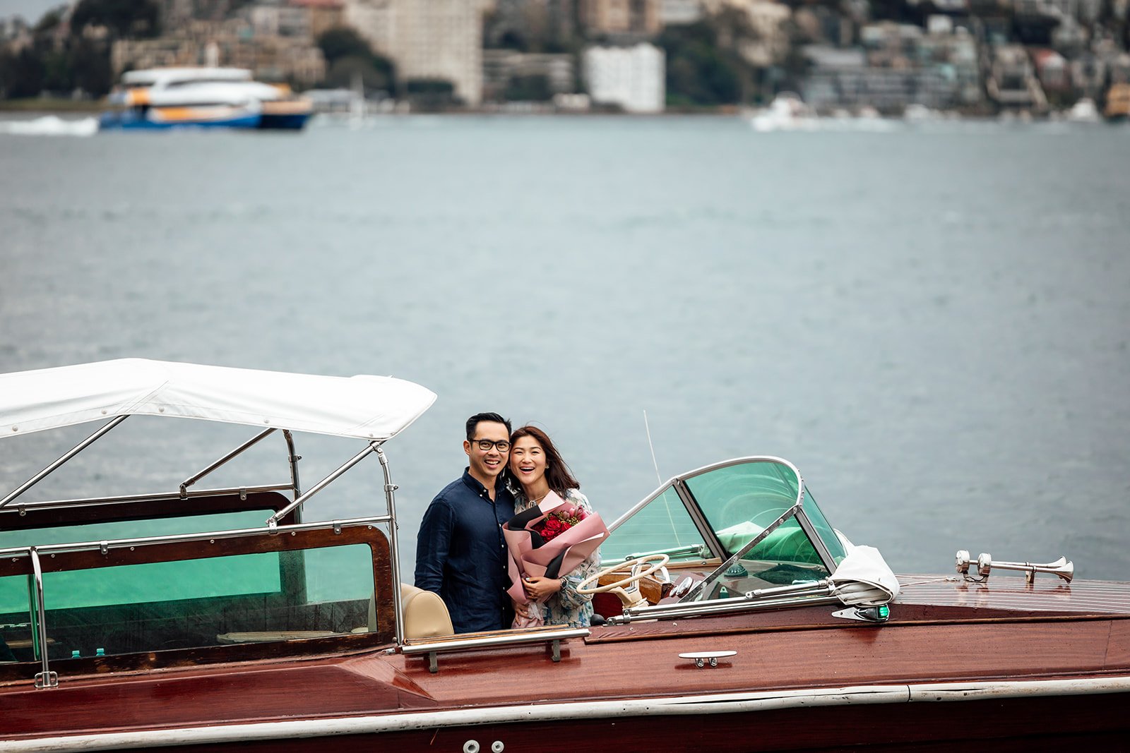 Couple on yacht holding flowers with Opera House in background.